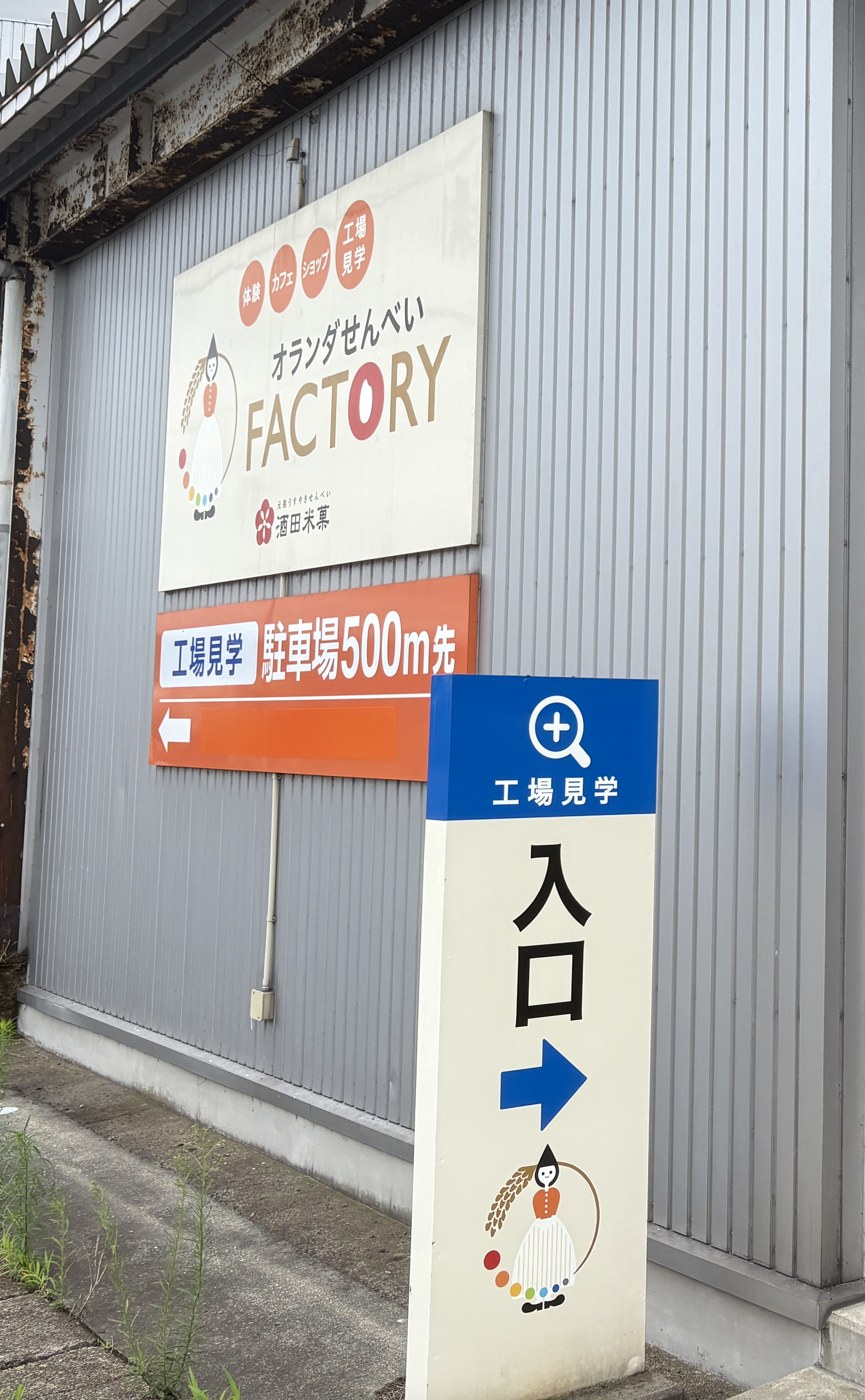 オランダせんべいFACTORYの壁に出た「工場見学 駐車場500m先」の看板と、「工場見学 入口→」の案内板